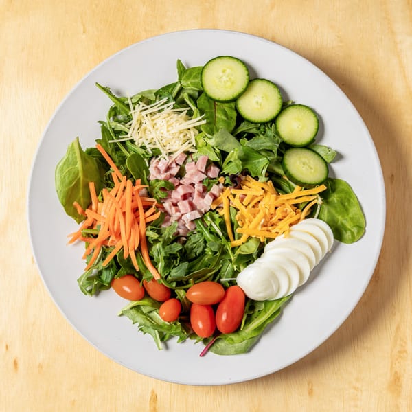 A colorful salad with greens, shredded carrots, ham, cheeses, cucumbers, tomatoes, and sliced eggs.